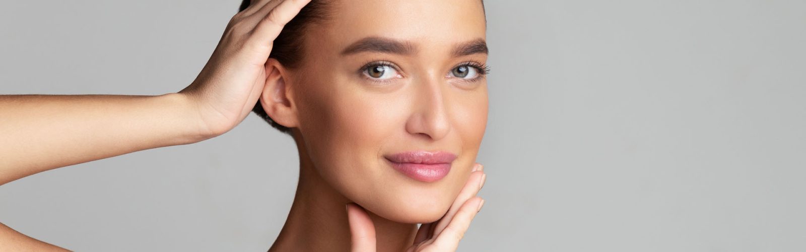 Beautiful woman touching face and looking at camera over grey background, free space, panorama